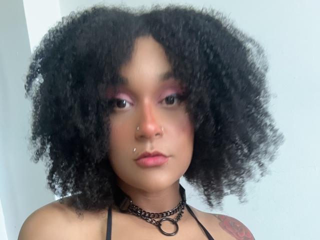 AmyStone69 with curly black hair, pink eye shadow, nose piercings, choker, and tattoo on right shoulder, looking straight at camera. Soft light highlights her smooth, brown skin.