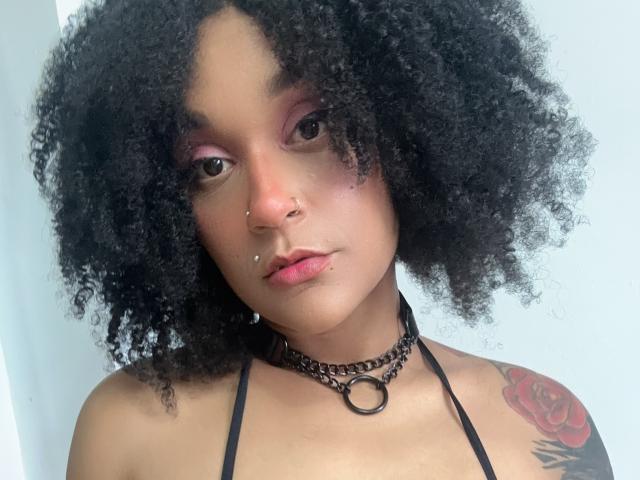 AmyStone69 with curly black hair, pink eyeshadow, nose piercings, lip piercings, black choker, tattooed red rose on right shoulder, wearing black halter top, looking at camera.