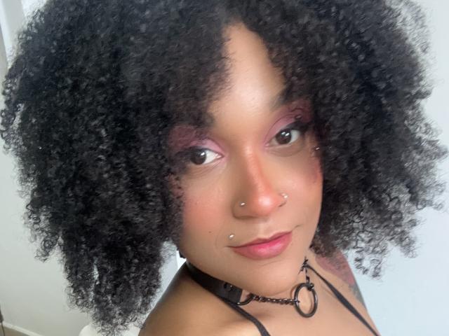 AmyStone69 with curly black hair, pink eyeshadow, nose piercings, lip piercings, and a black choker, smiling at the camera. She's got a cute tattoo on her left shoulder.
