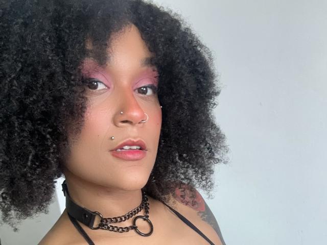 AmyStone69 with curly black hair, pink eyeshadow, nose piercings, lip piercings, black choker with metal ring, and tattoo on right shoulder, looking at camera with pink lips.