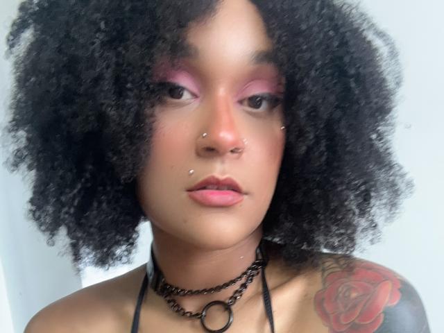 AmyStone69 with curly black hair, pink eyeshadow, nose piercings, lip piercings, choker, and a rose tattoo on her right shoulder. Her tits are not visible.