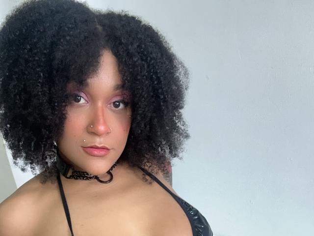 AmyStone69 has dark curly hair, a nose ring, purple eyeshadow, and a black choker with a heart ring. She's wearing a black strappy top, showing her tits.