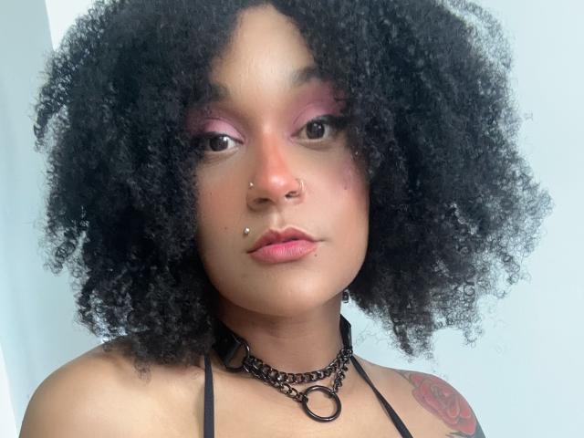 AmyStone69 with curly black hair, pink eye makeup, nose piercings, black choker, and tattoo on right shoulder, looking at camera with neutral expression.