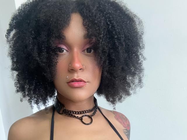 AmyStone69 with curly black hair, pink eyeshadow, nose piercings, lip piercing, black choker, ring, and tattoo on right shoulder, looking at camera with neutral expression.