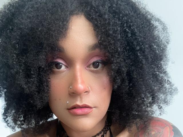 AmyStone69 has curly black hair, brown eyes, pink eyeshadow, nose piercings, and a tattoo on her right shoulder. She's wearing a black choker and has pink lipstick.