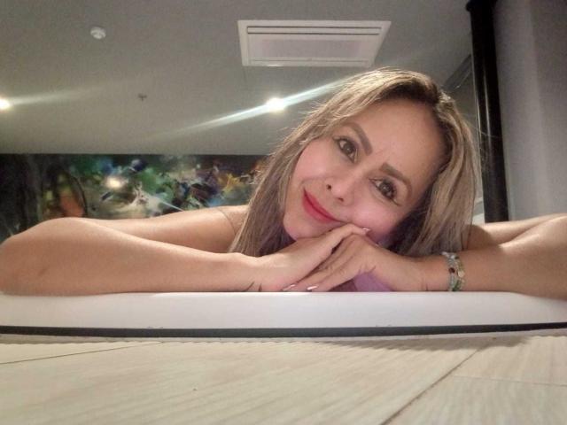 ScarletChanne mit blonden Haaren, rotem Lippenstift und einem süßen Lächeln, die ihr Kinn in die Hände legt und in einem Spiegel-Selfie ihre Brüste zeigt, trägt ein grünes Armband..