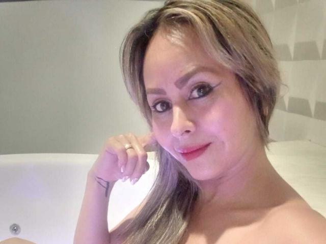 ScarletChanne mit kurzen, blonden Haaren, rotem Lippenstift und einem kleinen Tattoo am rechten Handgelenk, lächelt in die Kamera, die Hand ist in der Nähe ihres Gesichts, und sie zeigt ihre nackten Schultern..