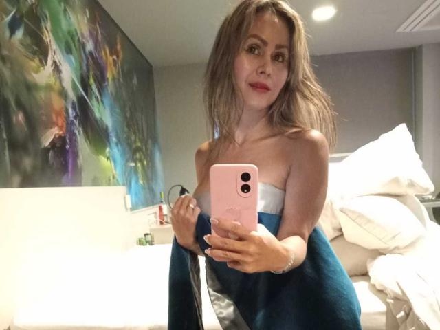 ScarletChanne macht ein Selfie in einem Schlafzimmer, trägt ein weißes Top ohne Träger und ein blaues Handtuch, hält ein rosa iPhone und hat eine hübsche, leicht gebräunte Brust und ein süßes Lächeln..