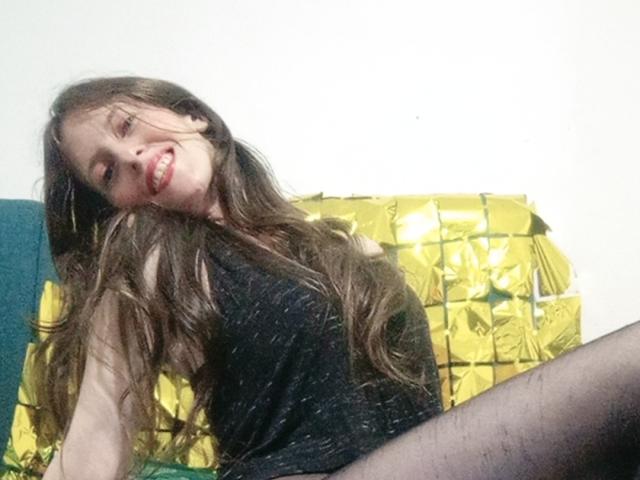 AlissonS69 has long brown hair, red lipstick, black sparkly dress, and black tights, sitting with a playful smile, yellow foil pillow in the background.