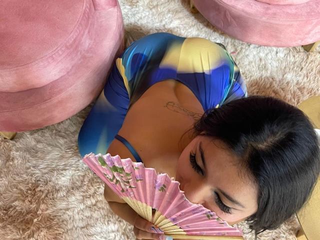 AlessiaFerry with dark hair, blue-yellow dress, holding pink fan, showing tits, tattoo on back, pink velvet chairs, lying on fluffy rug, eyelashes visible.