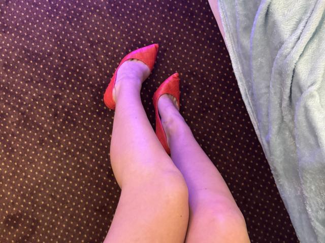 Close-up of NutaAlisson's legs in red pointy-toe shoes, standing on a brown carpet with small white dots. A light blue, textured fabric is on the right.