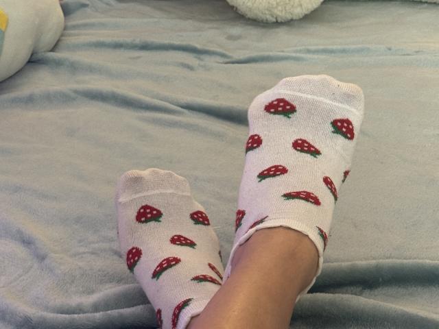 NutaAlisson's feet in white socks with red strawberry patterns, toes slightly curled, on a gray bed. Socks are slightly bunched at the ankles. Soft, textured fabric. Casual, relaxed.
