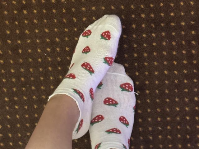 I piedi di NutaAlisson sono coperti da calzini bianchi con motivi di fragole rosse, leggermente sfilacciati ai bordi. Tappeto marrone con piccoli punti gialli sullo sfondo. I calzini sono incrociati..