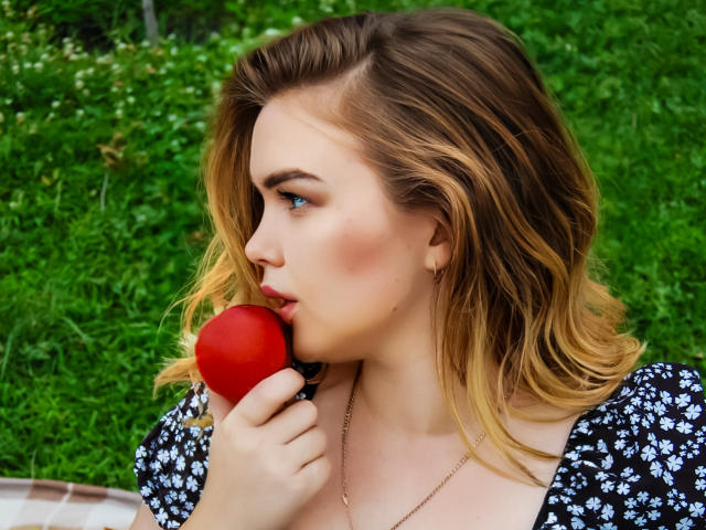 Kira Altman mit blonden Haaren, blauen Augen und geröteten Wangen, die einen roten Apfel beißt, trägt ein schwarzes Blumenkleid, eine goldene Halskette und Ohrringe, vor einem grünen, grasbewachsenen Hintergrund..