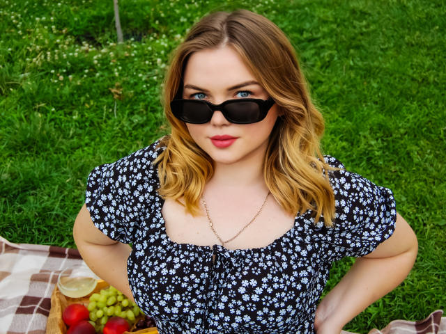 Kira Altman mit blauen Augen, die schwarze Sonnenbrille trägt, ein schwarzes Blumenkleid und eine goldene Halskette, steht auf grünem Gras, die Hände in den Hüften, blickt nach oben, und in der Nähe sind Trauben zu sehen..