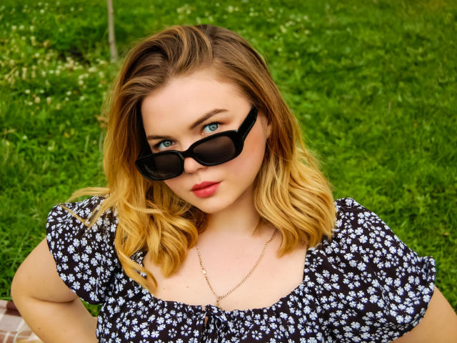 Kira Altman tem cabelo loiro e ondulado, olhos azuis, e usa óculos de sol pretos com um vestido floral preto e branco, e um colar de ouro. Seu batom vermelho se destaca..
