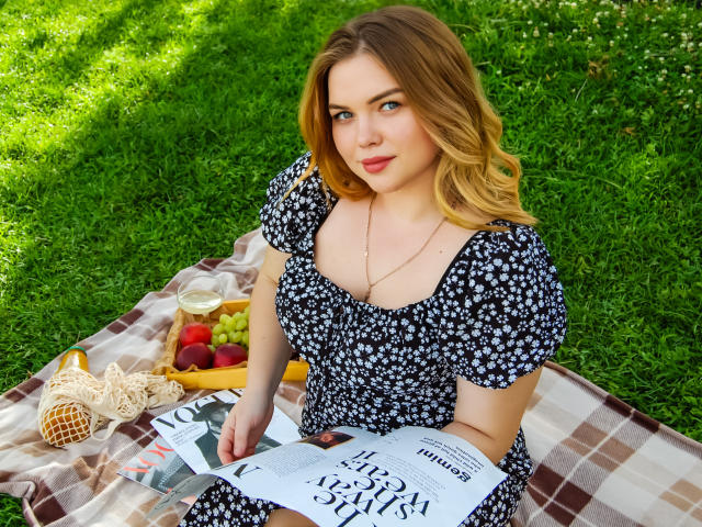 KiraAltman with blonde hair, blue eyes, small tits, wearing a black floral dress, reading a magazine on a checkered blanket, with a basket of fruit and a wicker bag.