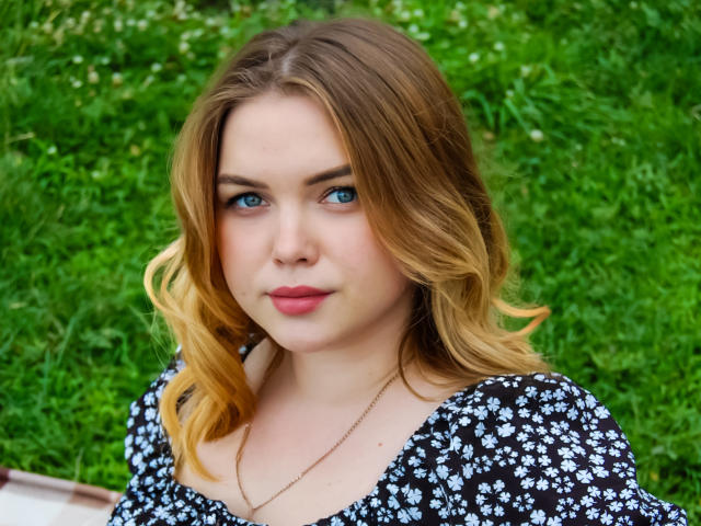Kira Altman, com cabelo loiro, olhos azuis e lábios rosados, vestindo um vestido floral preto, exibindo um leve sorriso, está em pé em frente a um gramado verde. Ela tem um traseiro atraente..