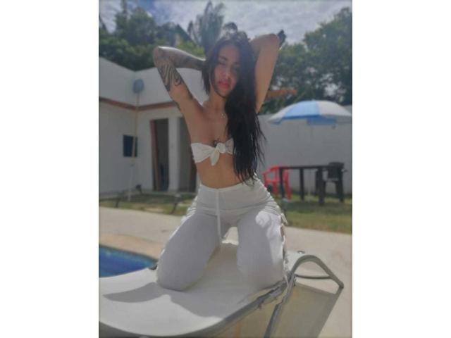 ErotiicAlexa with long black hair, white tied-up top exposing her tits, white pants, tattooed arms, kneeling on poolside table, sunny day, hands behind head.