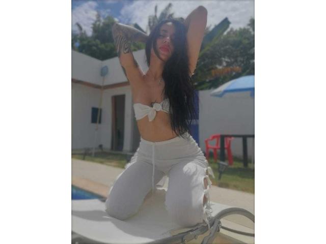 ErotiicAlexa with long black hair, white bikini top tied in front, showing small tits, white pants, kneeling on sunbed, arms raised, tattoos on left arm, outdoor, sunny.