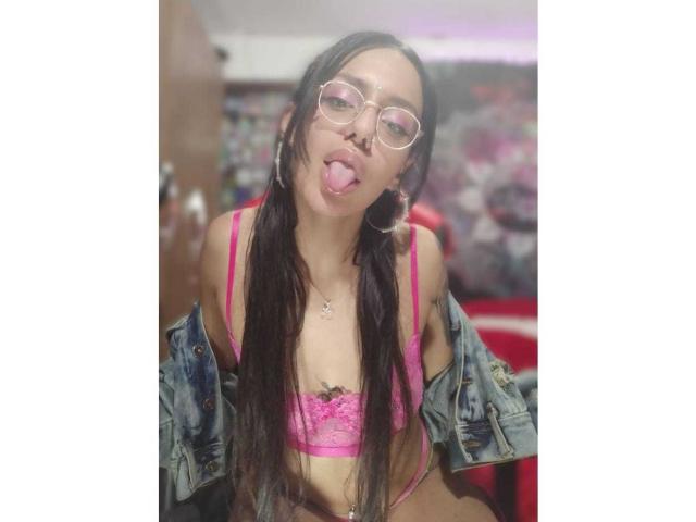 EroticAlexa with long black hair, glasses, and pink lace bra, tongue out, denim jacket off, showing small tits and a belly button piercing, in a slightly blurred photo.