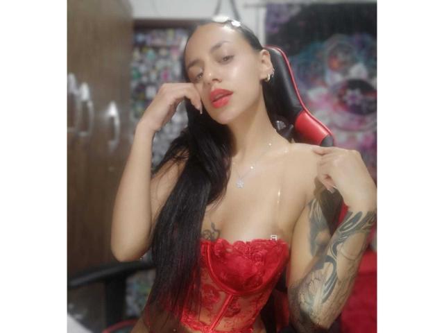 EroticAlexa with long black hair, red lace corset, and tattoos, sitting in a chair, hand on face, glossy skin, red lips, looking seductively at camera.