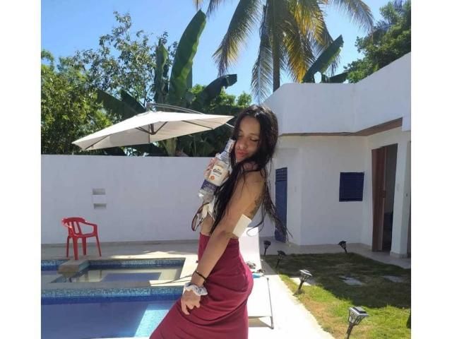 EroticAlexa with long black hair, red dress, and tan skin, holding a water bottle by a blue pool, under a white umbrella, with a red chair and greenery.