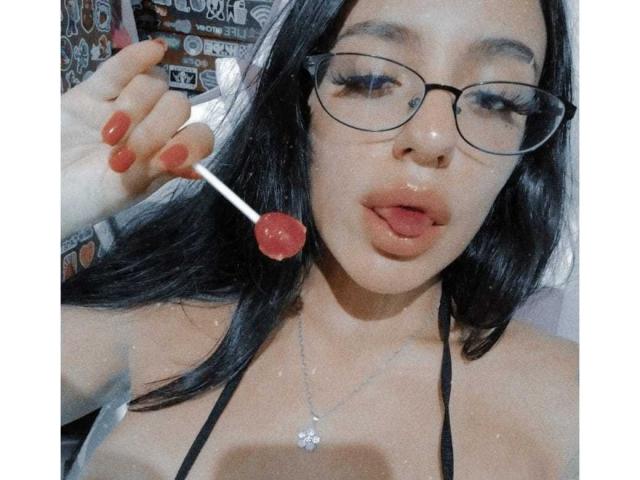 ErotiicAlexa with long black hair, glasses, and red nail polish, holding a red lollipop near her slightly open mouth, wearing a black halter top, showing her tits.