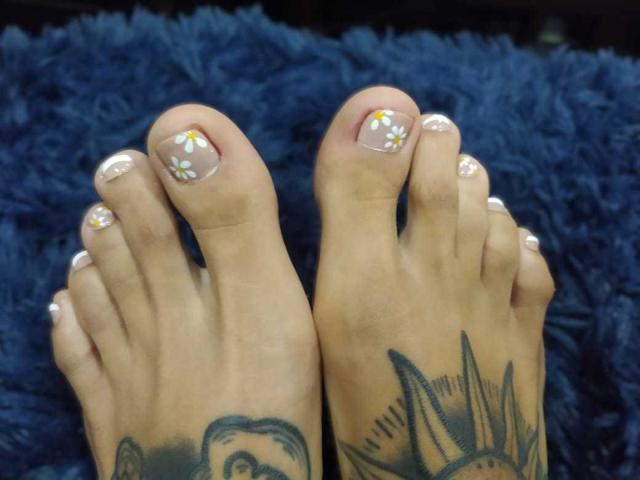 EroticAlexa's toes are painted with white nail polish and small yellow flowers, her feet have tattoos of a face and sun on the soles, on a blue textured surface.