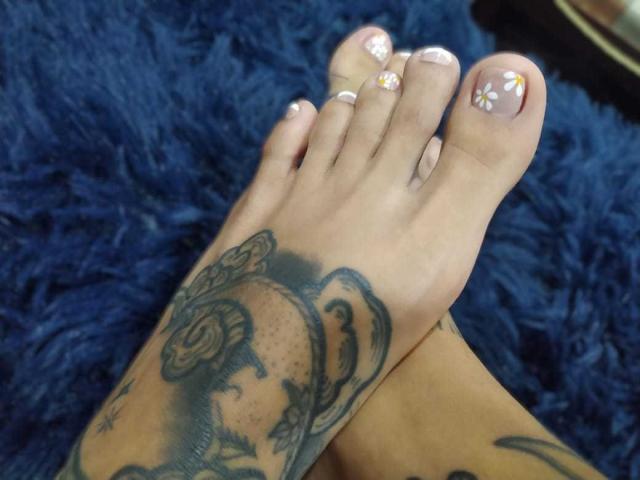 ErotiicAlexa's tattooed foot with a detailed black and grey portrait tattoo, crossed over her other foot. Her nails are painted with white and yellow floral designs.