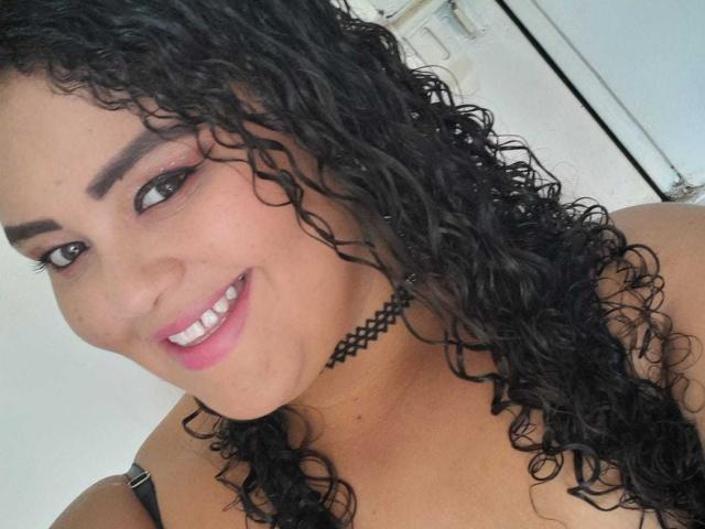 MelannieCarter has curly black hair, brown eyes, and a pink smile showing her white teeth. She's wearing a black choker and has a cute, attractive face with a sexy look.