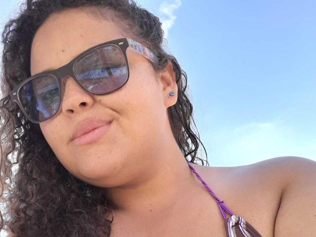 Melannie Carter tem cabelos pretos encaracolados, usa óculos de sol pretos e um top roxo de alças, com um pequeno brinco azul, e um leve semblante de descontentamento, sob um céu azul claro..