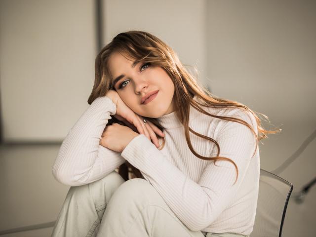 LauraHlot with long brown hair, wearing a white ribbed sweater, sitting with knees up, head resting on hand, looking at camera with a soft smile, in a clear chair.