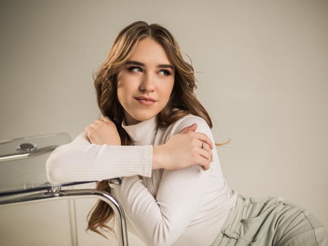 LauraHlot with wavy brown hair, white ribbed turtleneck, green pants, arms crossed, leaning on clear chair, soft smile, looking to the side, subtle makeup, relaxed pose.