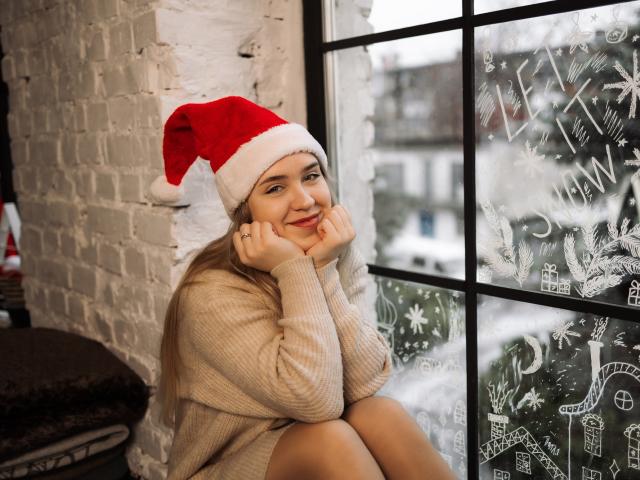 LauraHlot with long blonde hair, wearing a red Santa hat and beige sweater, sitting by a window with frosty patterns, hands on cheeks, smiling, legs crossed, butt slightly visible.