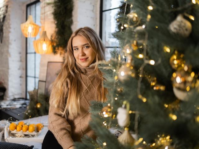 LauraHlot with long blonde hair, smiling, wearing a beige turtleneck, standing in front of a decorated Christmas tree, her tits slightly visible under her sweater, warm lights in background.