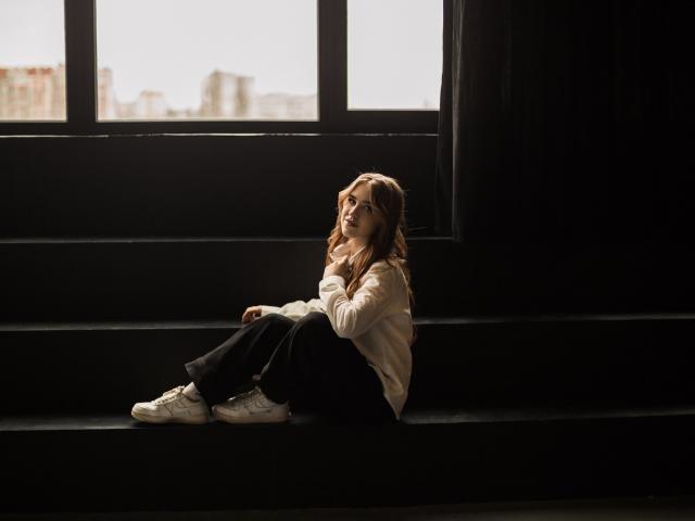 LauraHlot with brown hair, sitting on black bench, wearing white shirt, black pants, and white sneakers, looking up, light from window, soft shadows, relaxed, casual vibe.