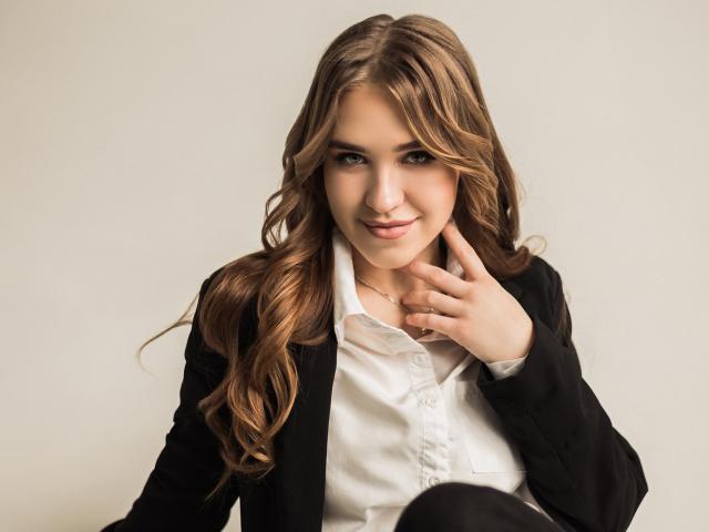 LauraHlot has wavy brown hair, green eyes, and a slight smile. She's wearing a black suit jacket over a white shirt, touching her neck with her right hand.