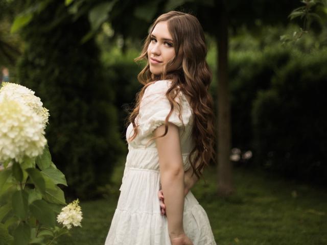 LauraHlot with long brown wavy hair, white dress, looking back, smiling, standing in green garden, white hydrangea flowers on left, soft natural light, slightly blurry background.