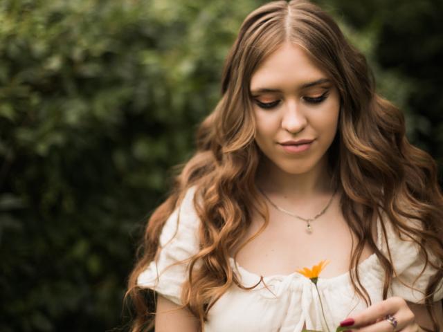 LauraHlot with long wavy brown hair, holding a yellow flower, wearing a white blouse, looking down, subtle smile, blurred green background, wearing a silver necklace.