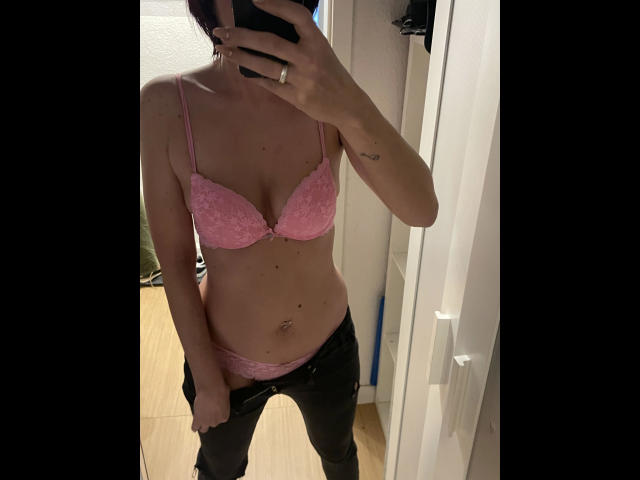 XxxLenaLustxxX-hot in pink lace bra and panties, black jeans unbuttoned, holding phone mirror selfie, showing tits and a belly button piercing. Tattoo on right forearm.