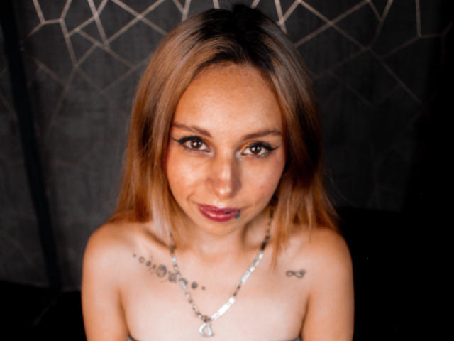 KateBrunet with light brown hair, looking up, has tattoos on her chest, black eyeliner, and a silver necklace, topless, showing her small tits, and a subtle smile.