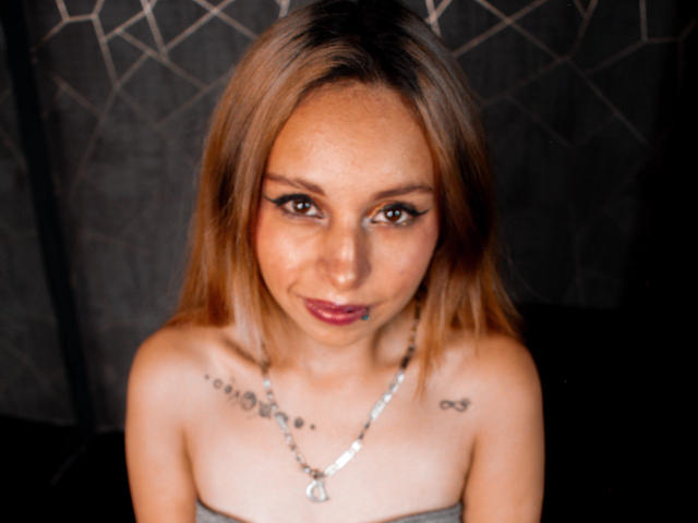KateBrunet has light brown hair, dark eyeliner, and red lipstick. She's topless with small tits, a necklace, tattoos on her chest, and a slight smile.