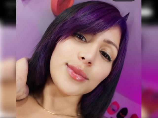 RaquelZ has purple and black hair, dark eyes, glossy pink lips, and a subtle smile. She's wearing a gold necklace with her arm behind her head.