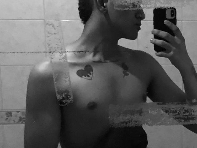 Black-and-white mirror selfie of Dekk, shirtless, showing a heart tattoo on left chest and a small bird tattoo on right chest, holding a phone, with wet glass.
