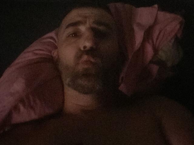 LartisteBabes, bald with a beard, lying in bed, shirtless, pink pillow under head, dark room, serious expression, slightly blurry, brown skin, dark hair, dark eyes, visible chest.