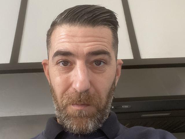 LartisteBabes has a bearded face with dark, slicked-back hair, brown eyes, and a serious expression, wearing a dark blue shirt. His nose is slightly upturned.