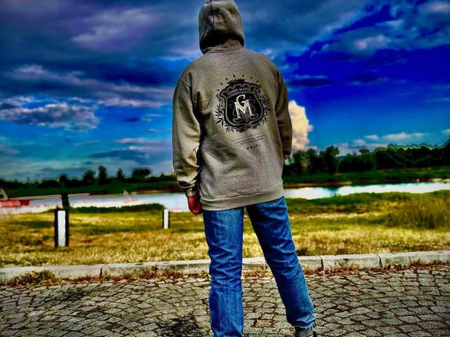 WarzyWkawcipce stands with back to camera, wearing a gray hoodie and blue jeans, under a vivid blue sky. Hood up, hood art visible, standing on cracked pavement.