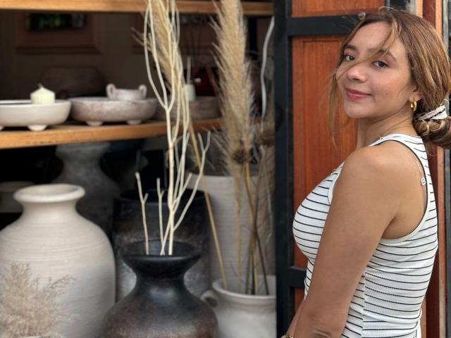 MajoGomez with small tits, wearing a white striped tank top, smiling, standing beside pottery and dried reeds, has brown hair in a messy bun, and a cute face.