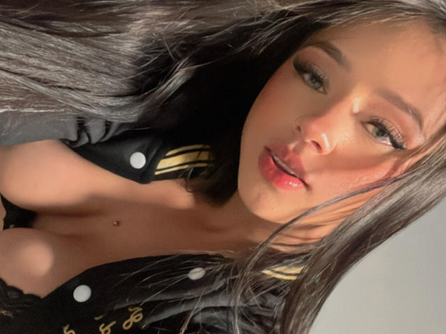Kloe Thompson, con lunghi capelli neri, indossa una giacca da aviatore nera, mostrando il seno, con un piccolo piercing all'ombelico e labbra piene di colore rosa, mentre guarda nella fotocamera..